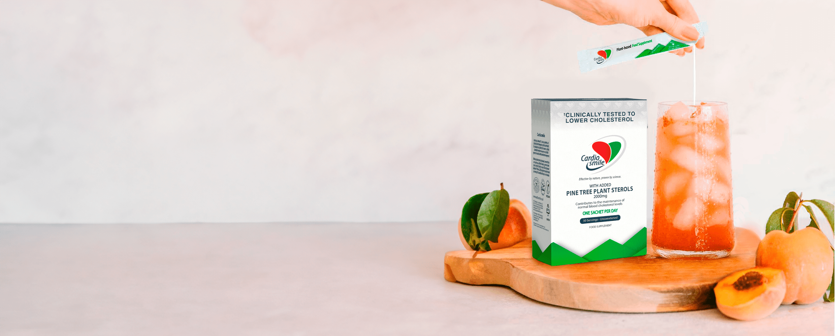 CardioSmile plant sterols box on a wooden board, with a hand pouring a sachet into an iced pink drink beside peaches.