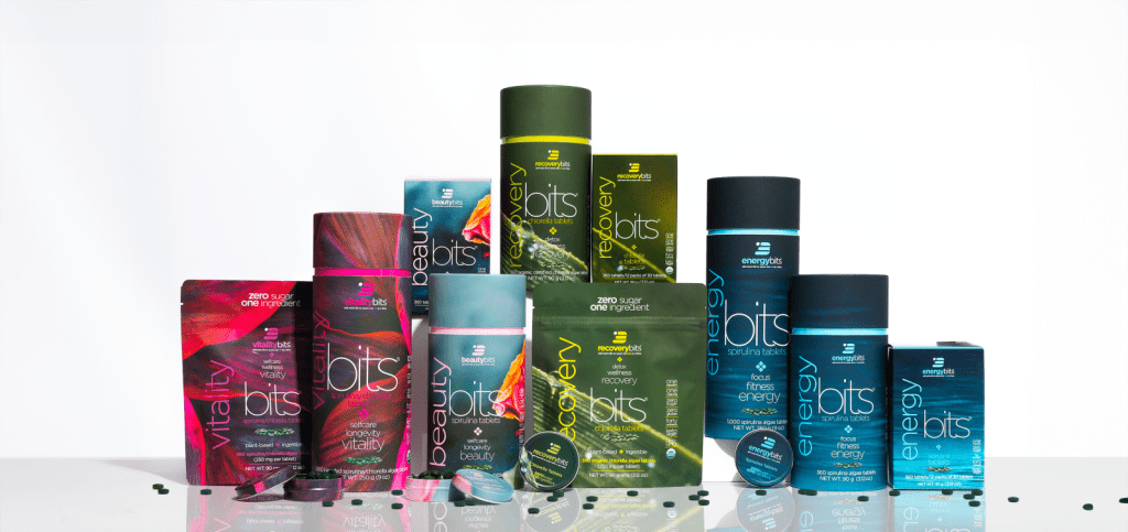 ENERGYbits product family arranged on a white reflective surface—canisters, pouches, and tins of ENERGYbits (spirulina), RECOVERYbits (chlorella), BEAUTYbits, and VITALITYbits—with a few dark green tablets scattered in front.
