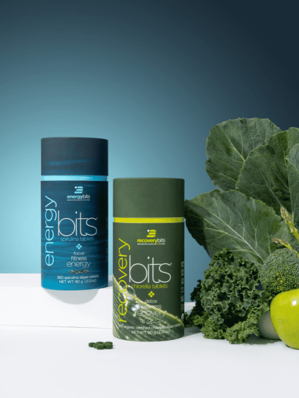 ENERGYbits canisters—ENERGYbits (spirulina) and RECOVERYbits (chlorella)—on a white surface with kale, broccoli, and a green apple; teal gradient background and a few tablets in front.